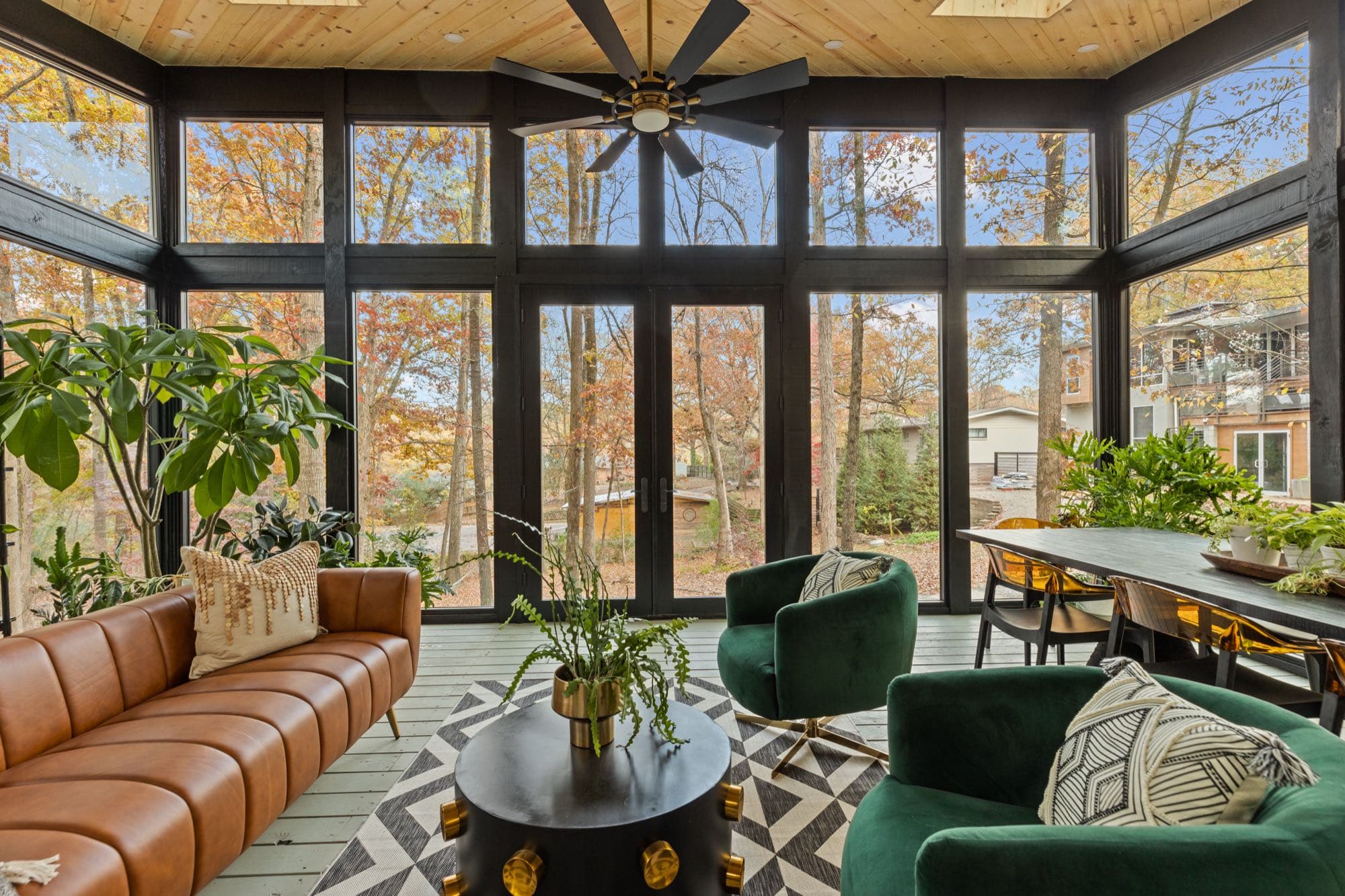 Glass walled sunroom in a short term rental, with a leather sofa, velvet chairs, and fall woods and a neighboring house beyond the glass