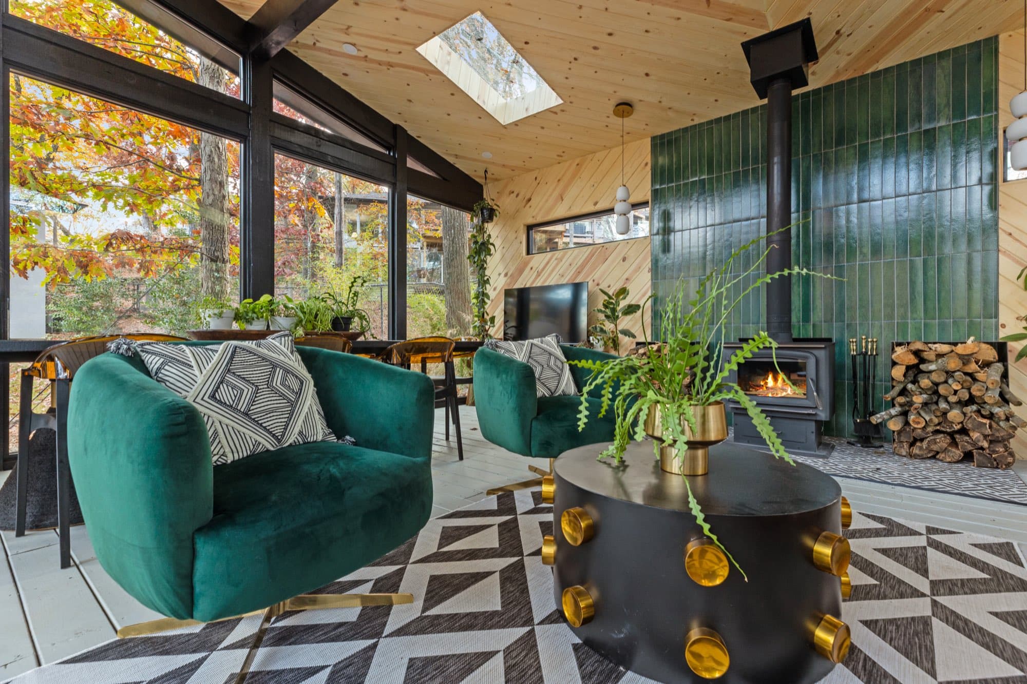 Short term rental sunroom, with emerald velvet chairs beside a lit wood stove on a glossy green tile wall and firewood stacked alongside