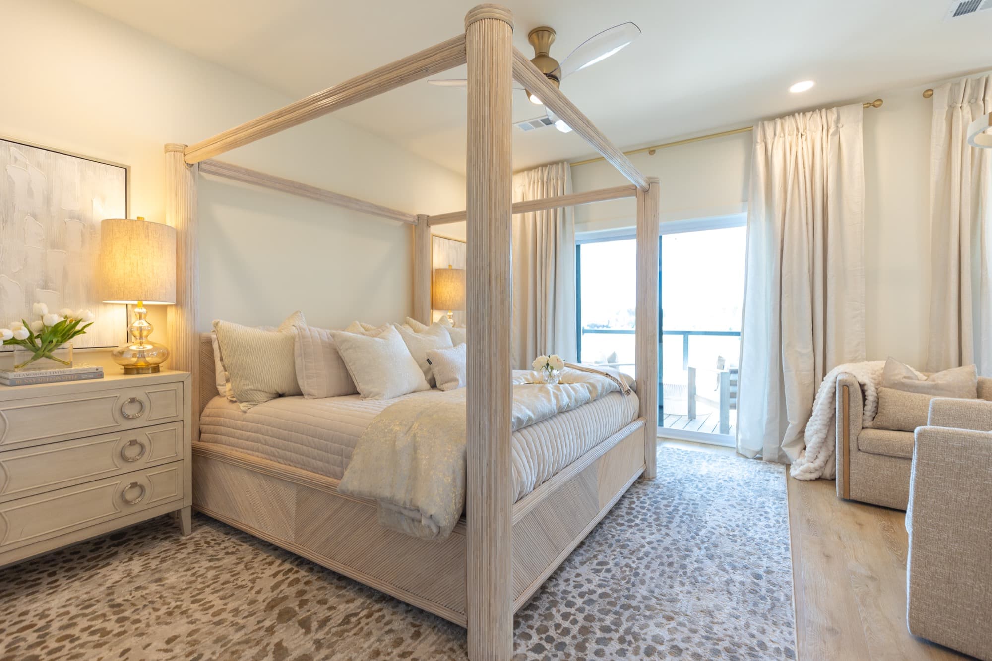 Primary bedroom in a short term rental, with a cane canopy bed, layered cream bedding and a glass balcony door behind full length drapes