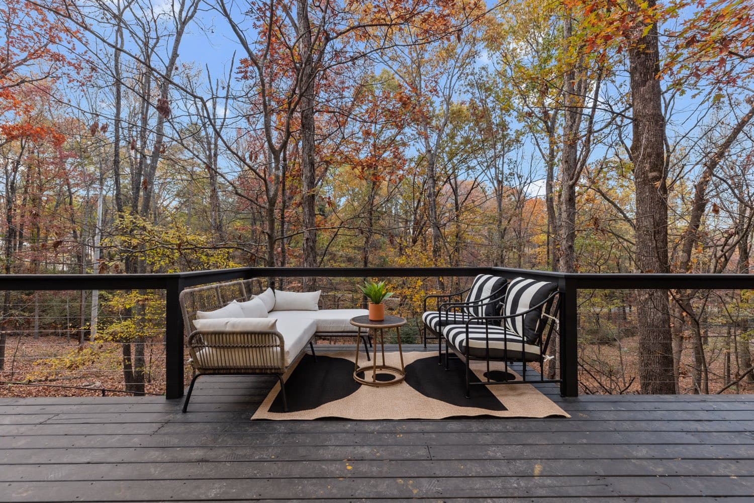 Deck of the stilt cabin rental, with a rope daybed and striped chairs behind cable railing, under a thinning oak canopy and open sky