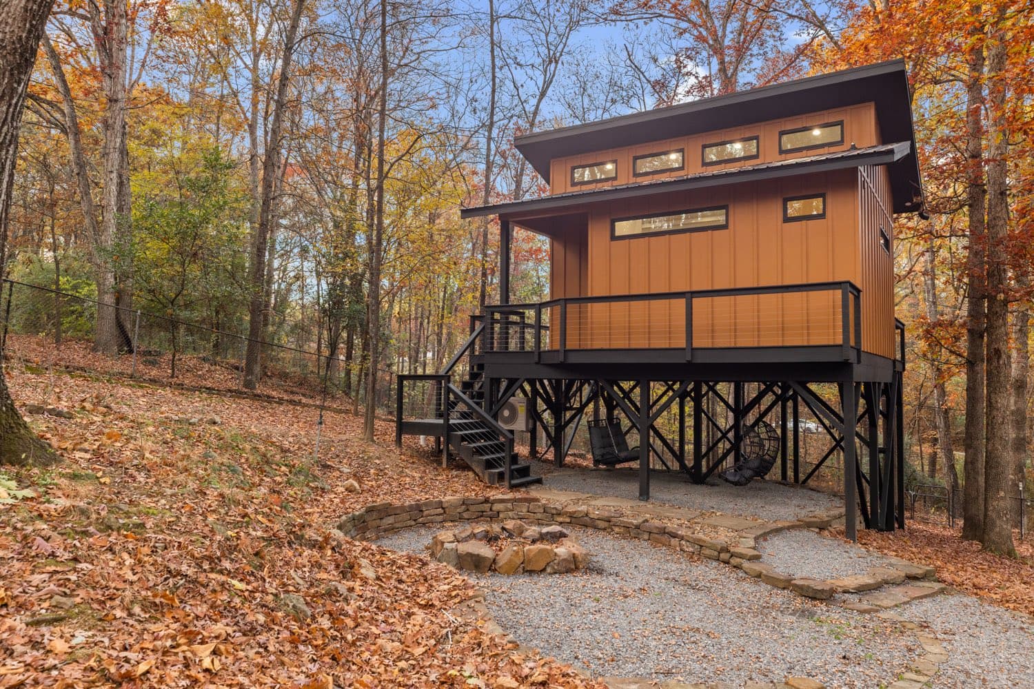 Modern short term rental cabin on black steel stilts in a fall oak forest, with a cable rail deck and a stone fire pit