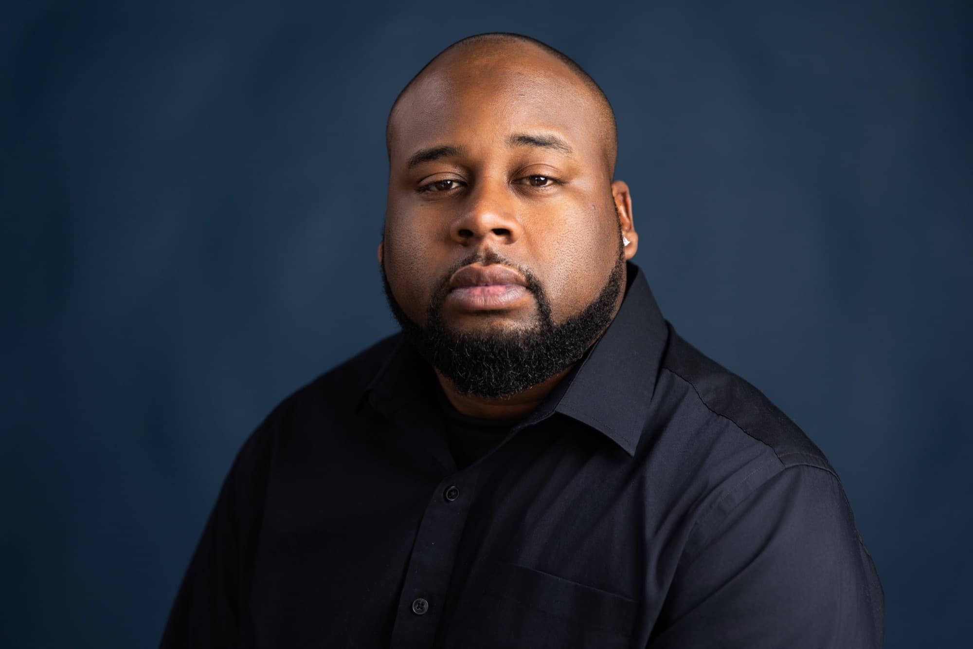 A man with a shaved head and dark beard in a black shirt, looking straight at the camera against a mottled deep blue backdrop