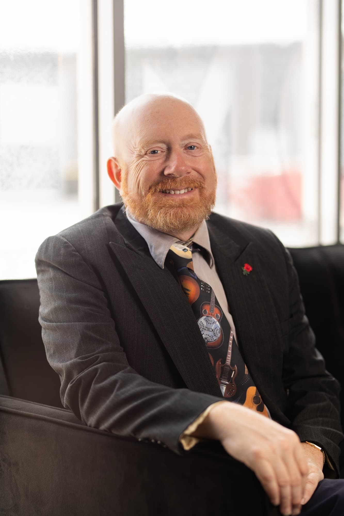The same man back in the guitar print tie, seated with one arm along dark velvet upholstery, in front of a bright daylit window