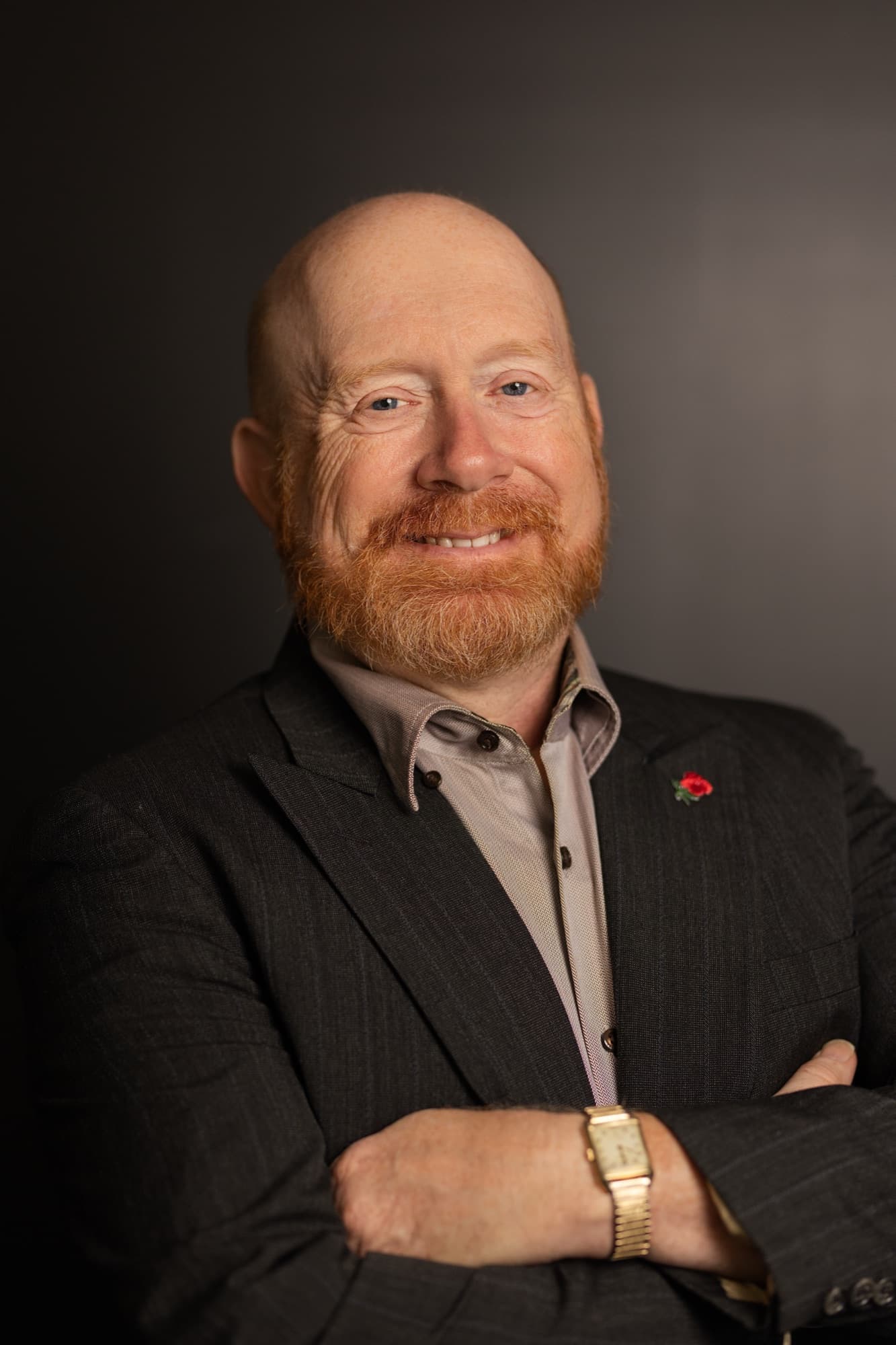 The same man in the same pinstripe jacket, tie removed and taupe shirt collar open, arms folded, against the dark gray backdrop