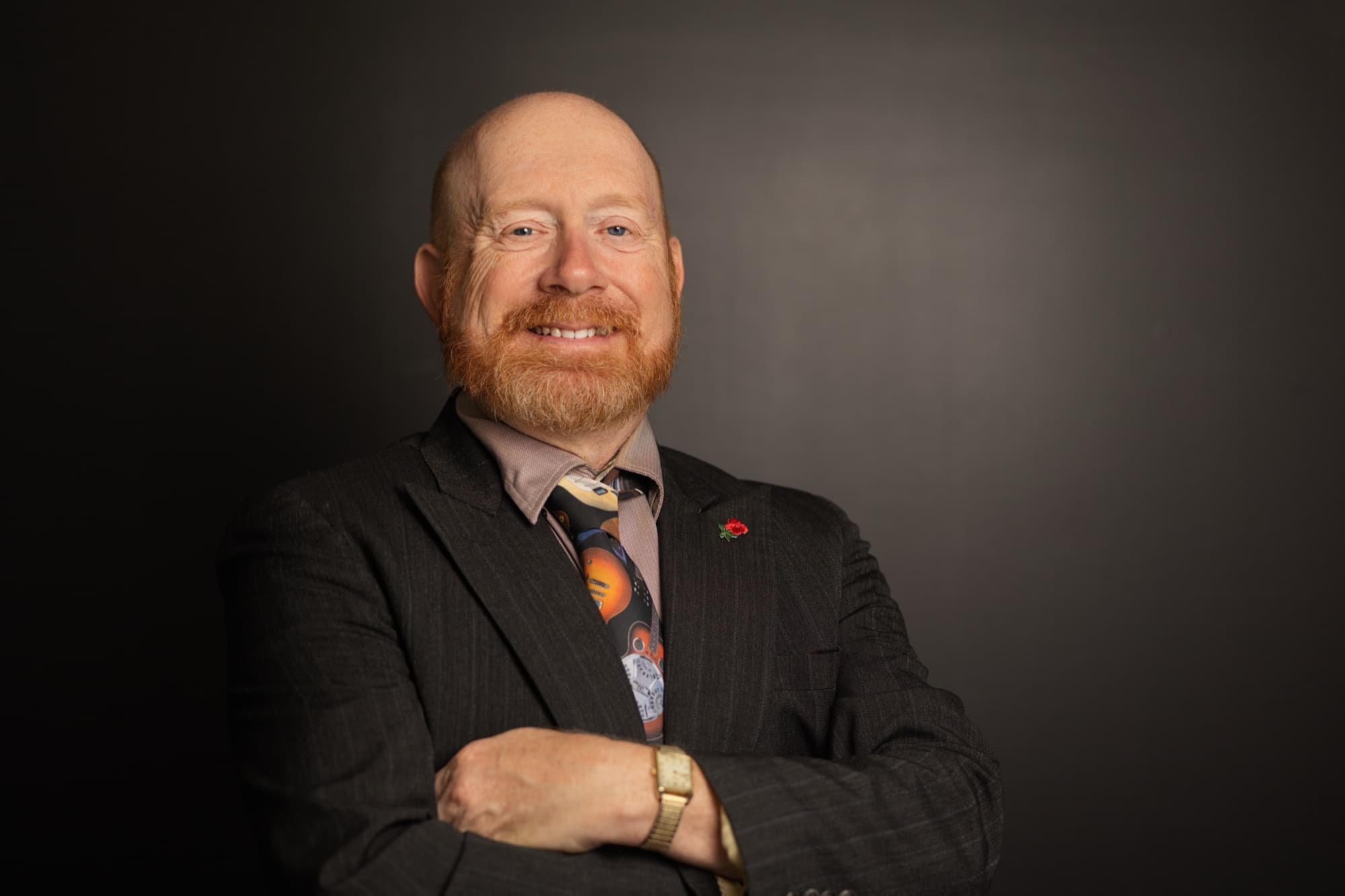 A bald man with a red beard in a charcoal pinstripe jacket and a guitar print tie, arms folded, against a dark gray studio backdrop