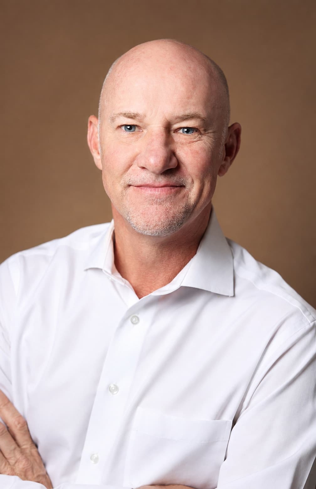 A bald man with gray stubble in a crisp white dress shirt, head and shoulders, against a warm brown studio backdrop