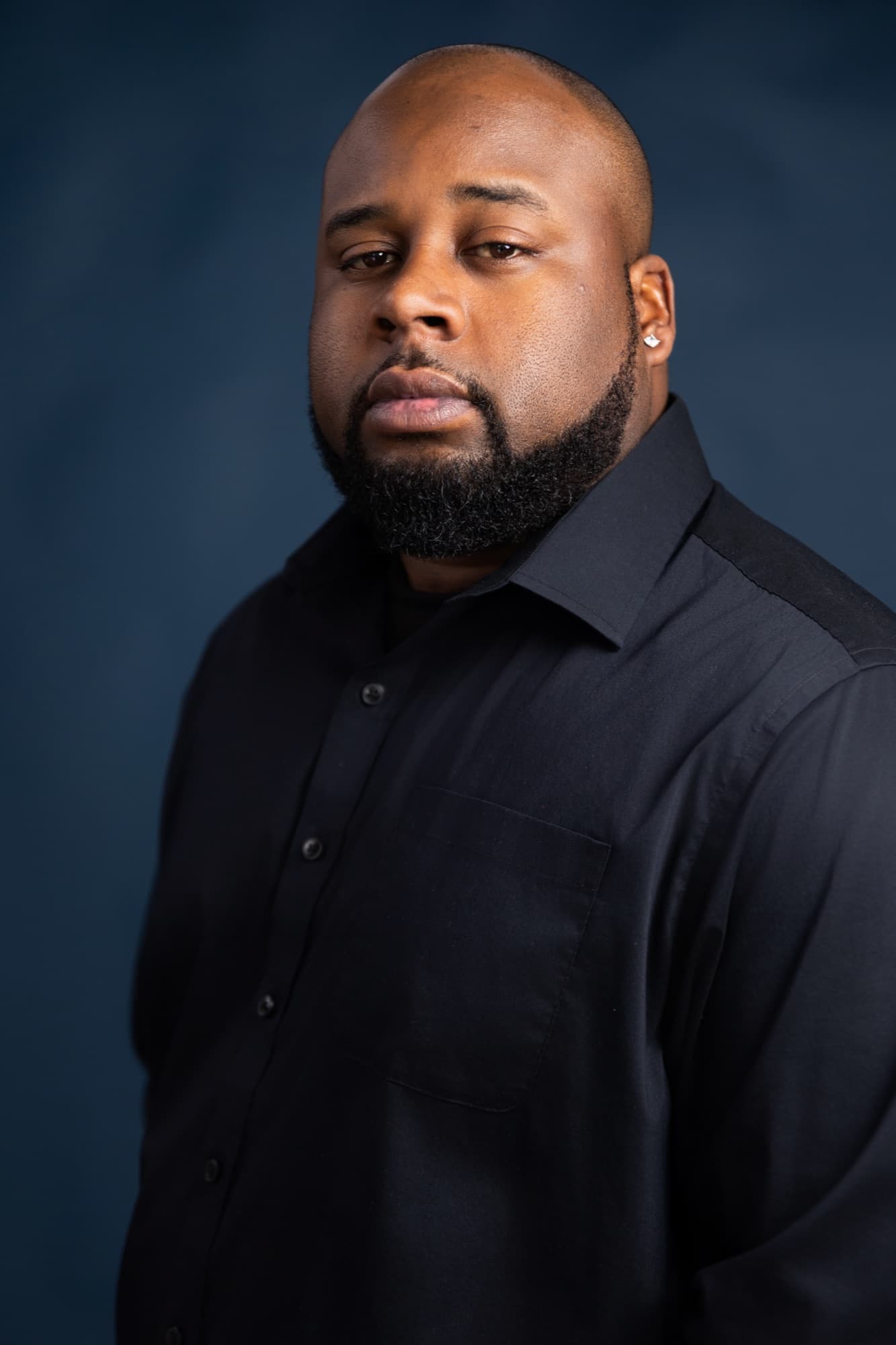 A man with a shaved head and dark beard in a black button down shirt, turned to one side against a mottled deep blue backdrop