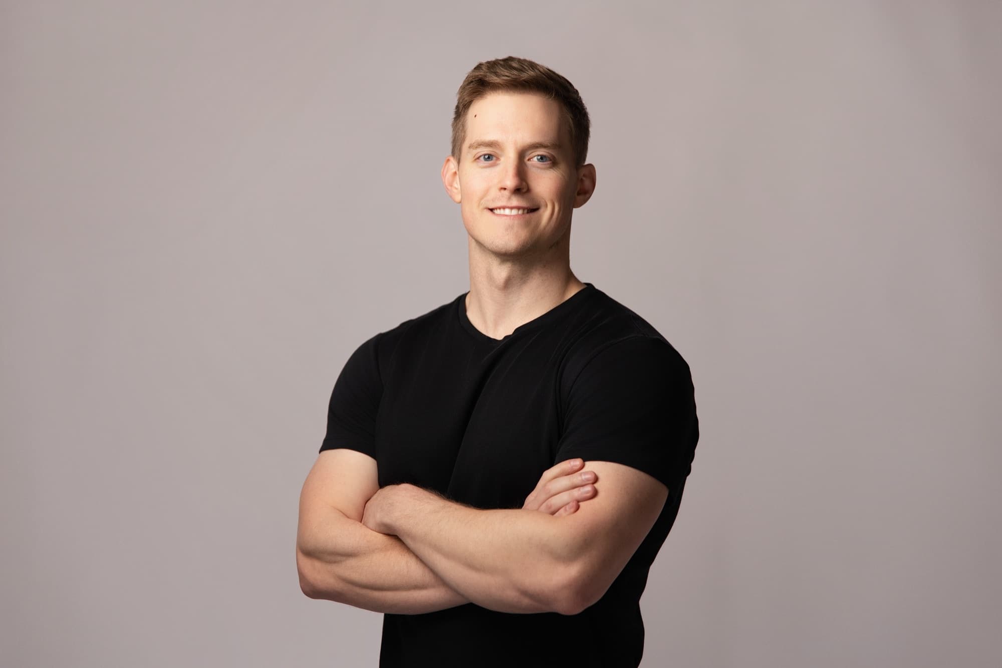 A man with short mid brown hair in a plain black t-shirt, arms folded, with a slight smile, against a warm gray seamless backdrop
