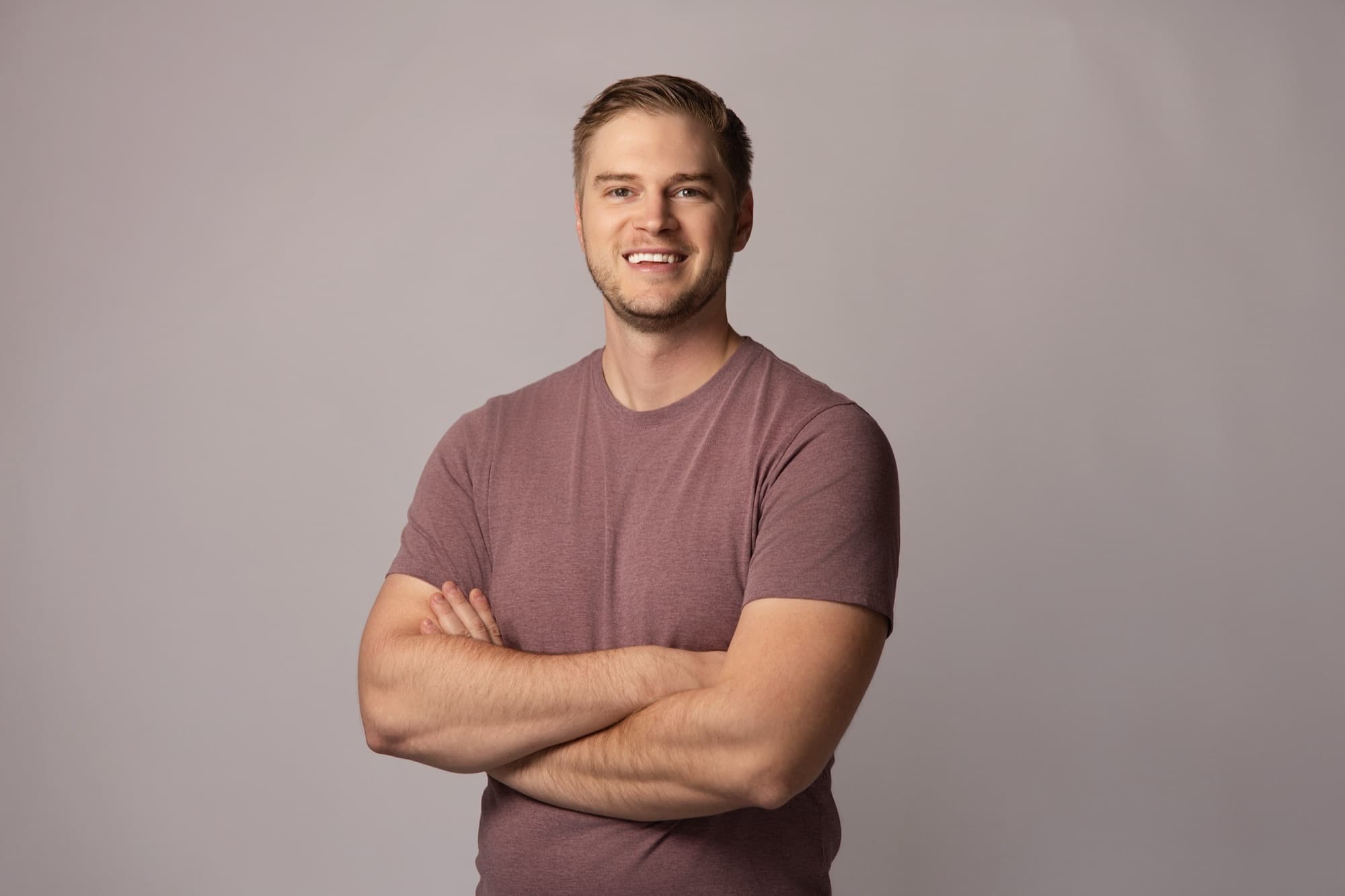 A man with short fair hair in a heathered mauve t-shirt, arms folded and smiling broadly, against a warm gray seamless studio backdrop
