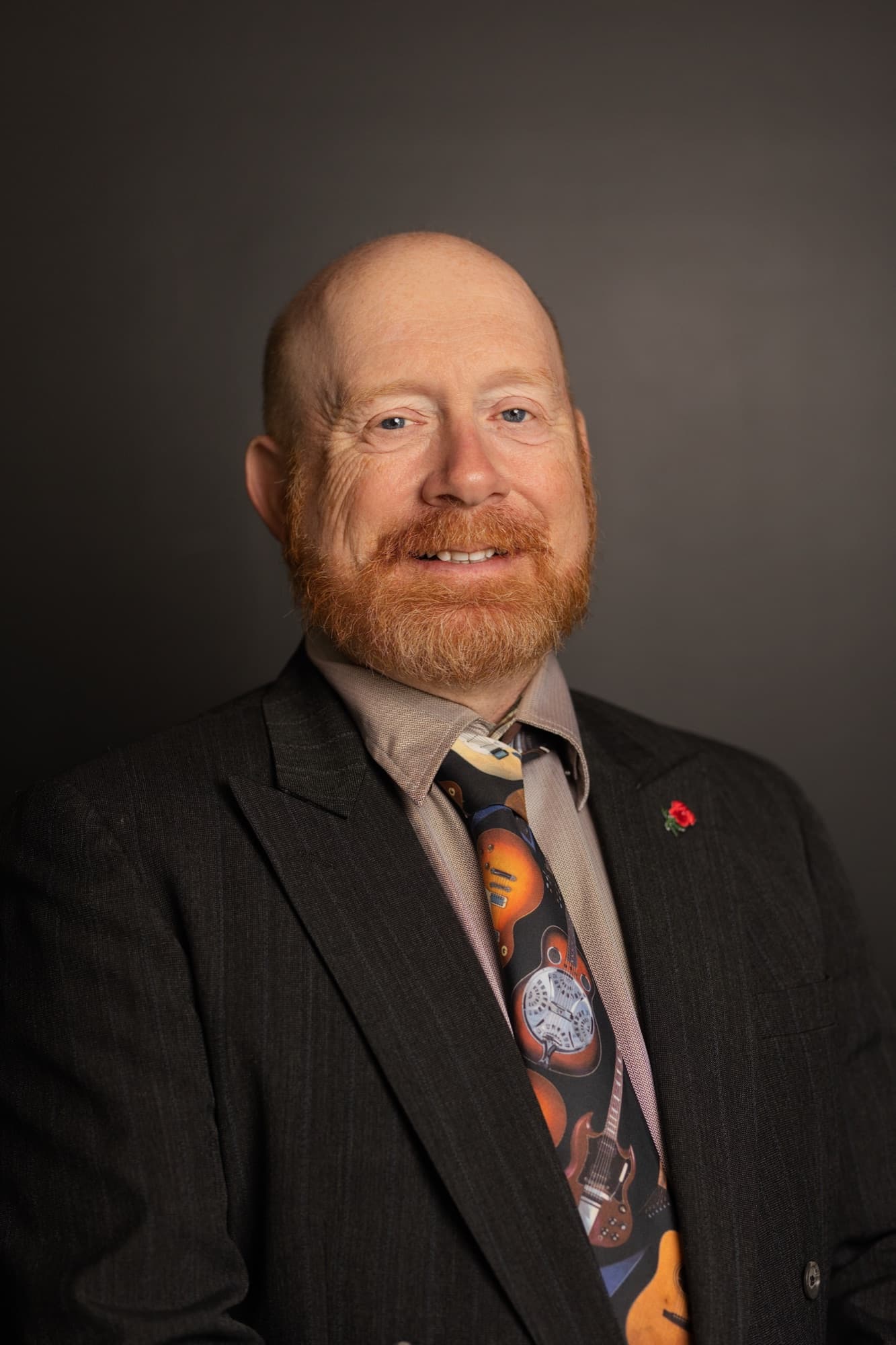 A bald man with a red beard in a charcoal pinstripe jacket, taupe shirt and a guitar print tie, smiling against a dark gray backdrop