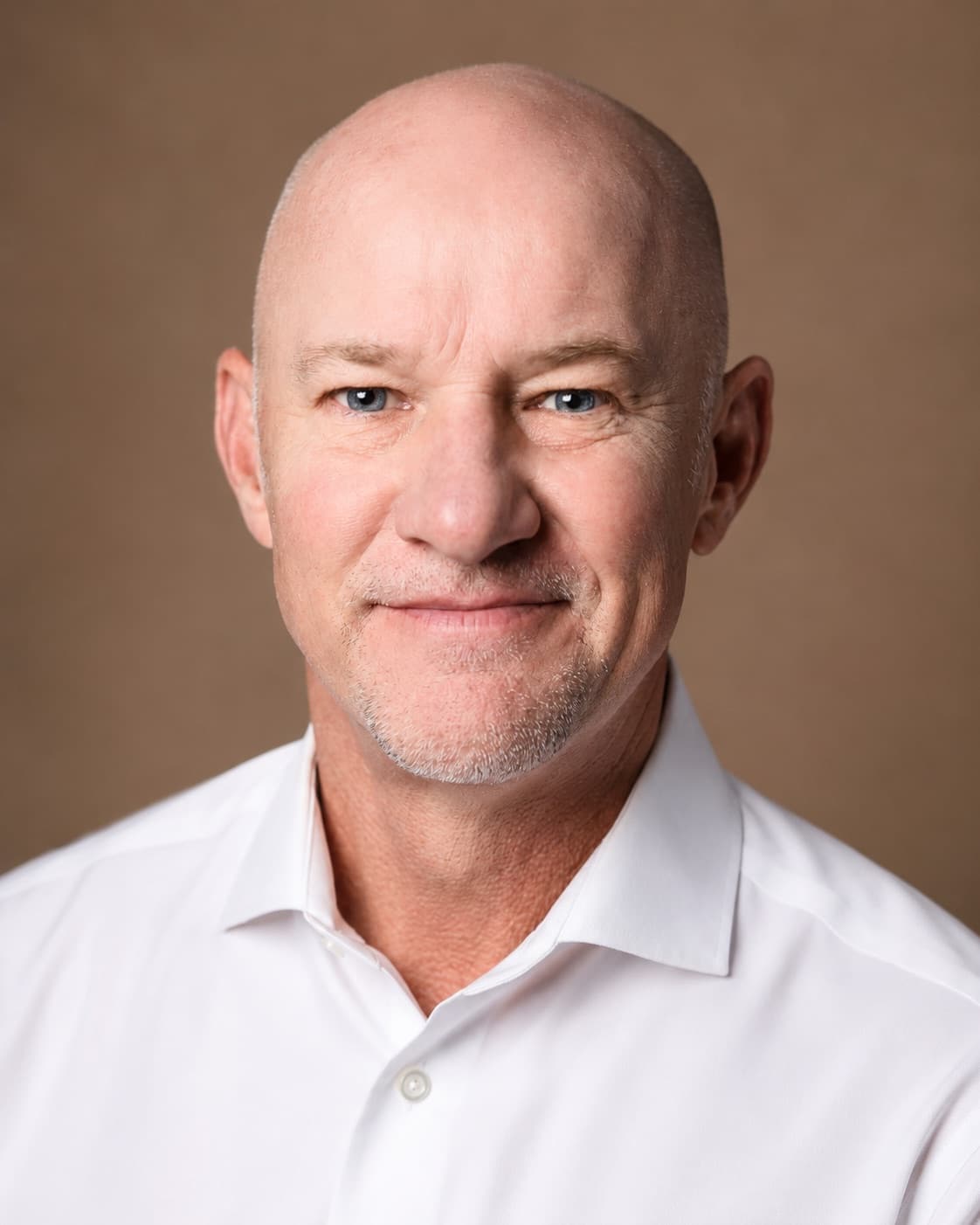 A bald man with gray chin stubble in a white open collar dress shirt, smiling with lips closed, against a mottled warm brown backdrop