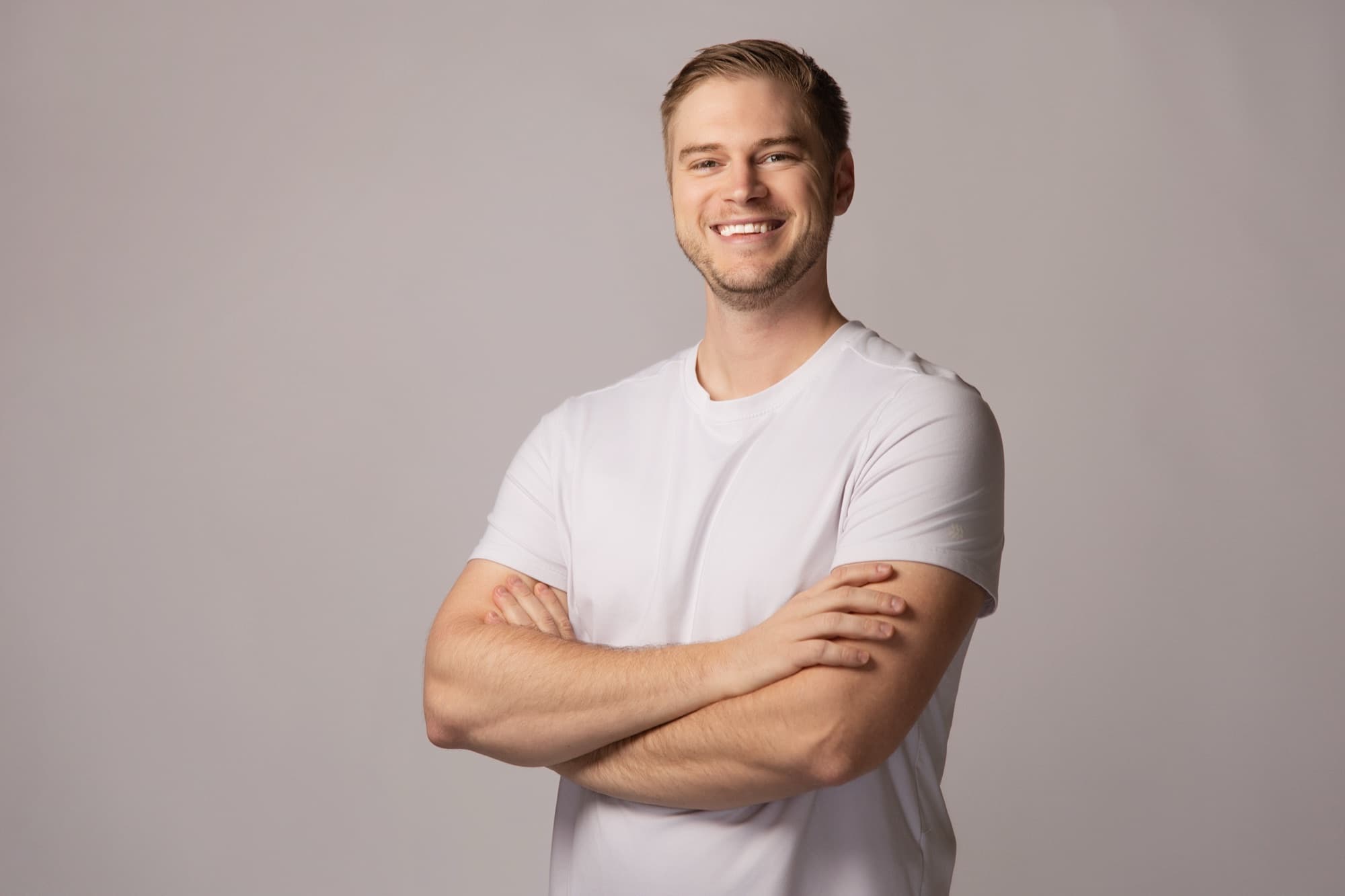 A younger man with short fair hair in a plain white t-shirt, arms folded, smiling broadly against a warm gray seamless backdrop