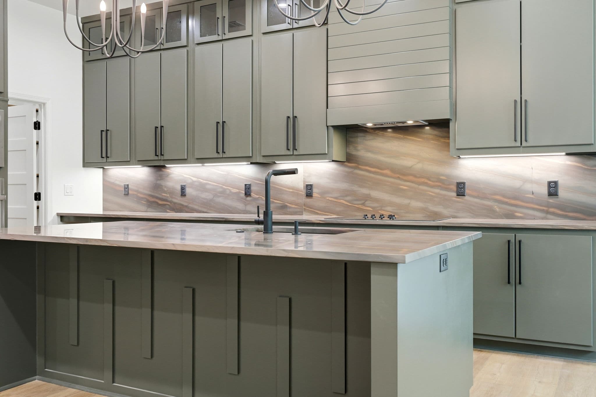 Kitchen island with a sink, sage green cabinets and a veined stone backsplash in a new construction home in Cabot, Arkansas