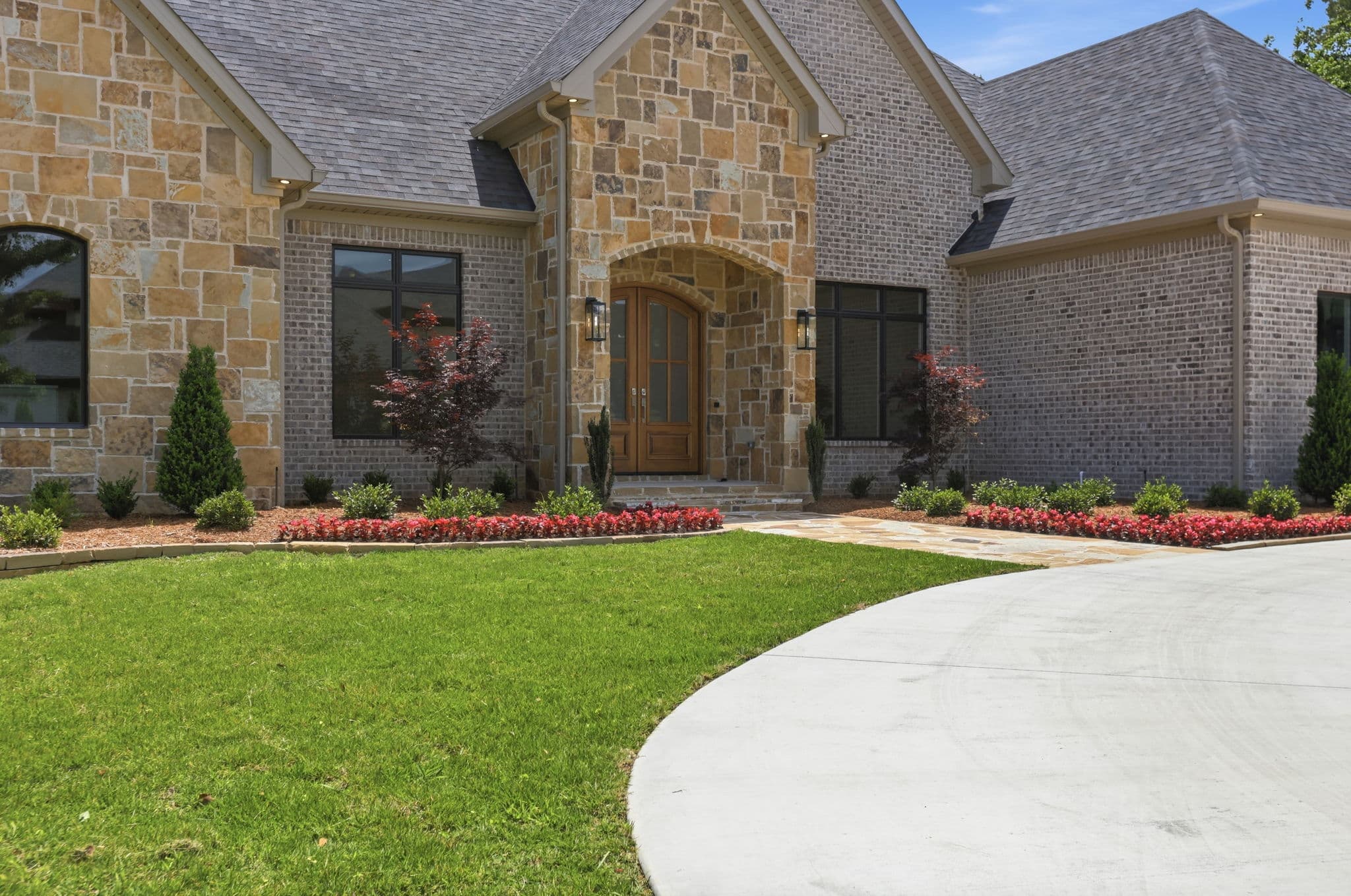 Stone and brick French country home in Little Rock, Arkansas, photographed in bright daylight under a blue sky, with finished beds, a green lawn and a curved concrete driveway