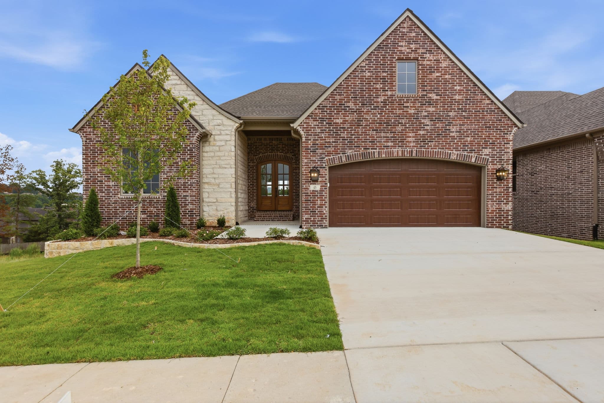 Finished brick and stone front elevation of a new construction home in Little Rock, Arkansas, with fresh sod, a young staked tree and a brown double garage door