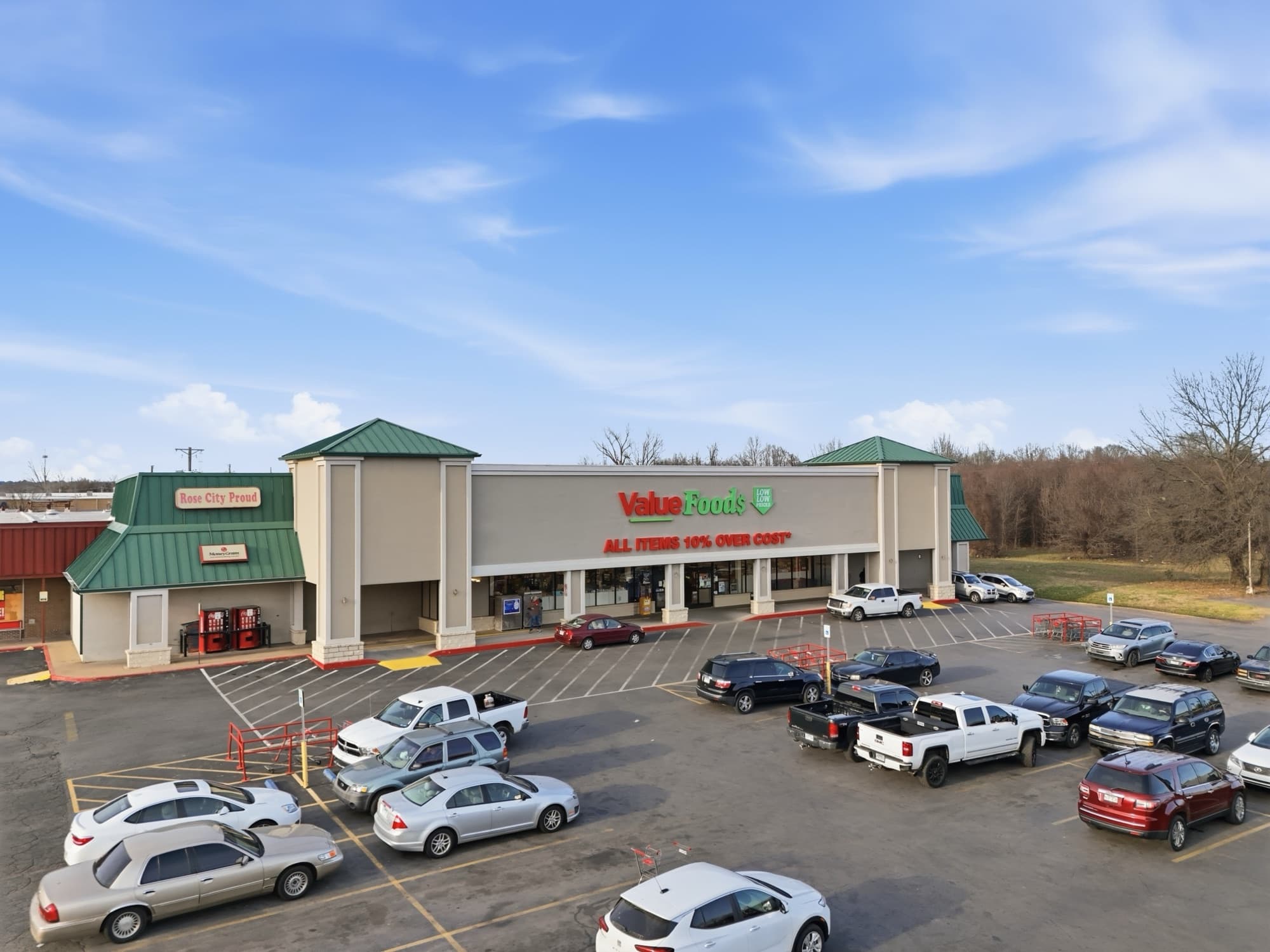 Elevated three quarter view of the grocery anchor at Rose City Center in North Little Rock, Arkansas, with a tan stucco facade, green standing seam gables and a parking lot of cars and pickups under a blue sky