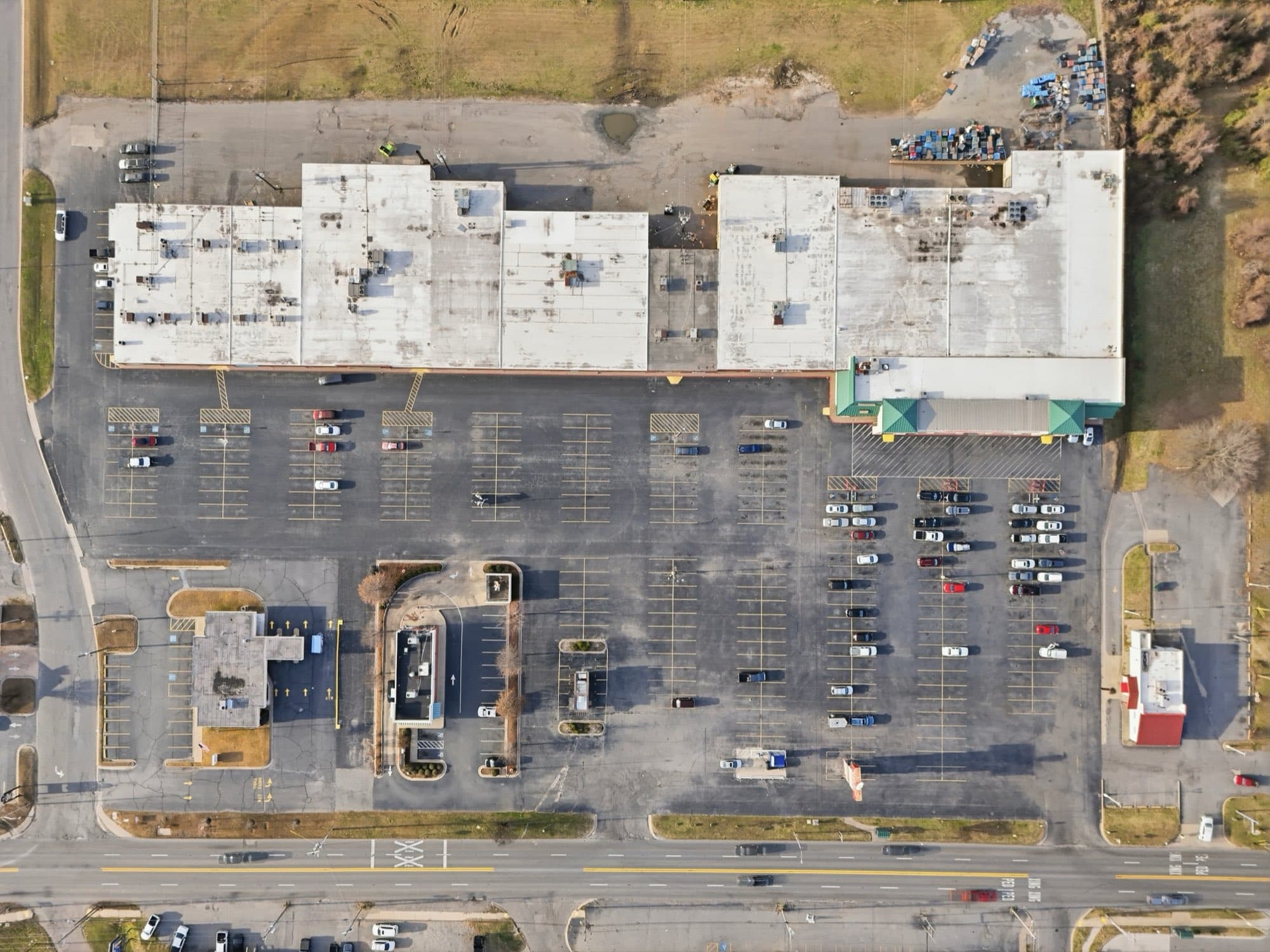 Overhead drone photo of Rose City Center in North Little Rock, Arkansas, showing the white flat roofs of the strip and its grocery anchor, the full striped parking field, an out parcel building with a drive through lane, and a four lane highway along the bottom of the frame
