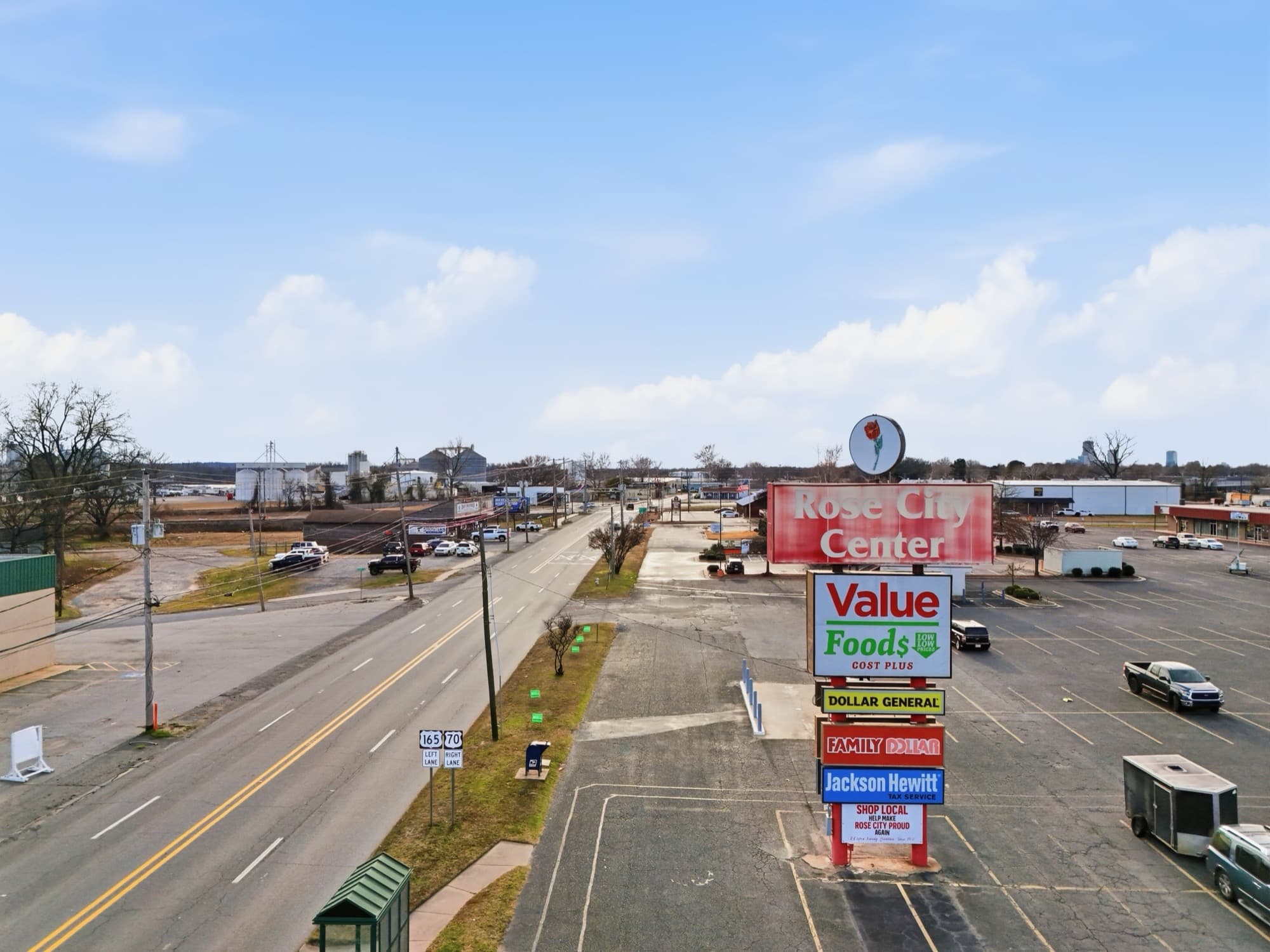 Elevated view of the Rose City Center pylon sign in North Little Rock, Arkansas, with a rose logo above stacked tenant panels, beside a highway with route markers and grain silos on the horizon