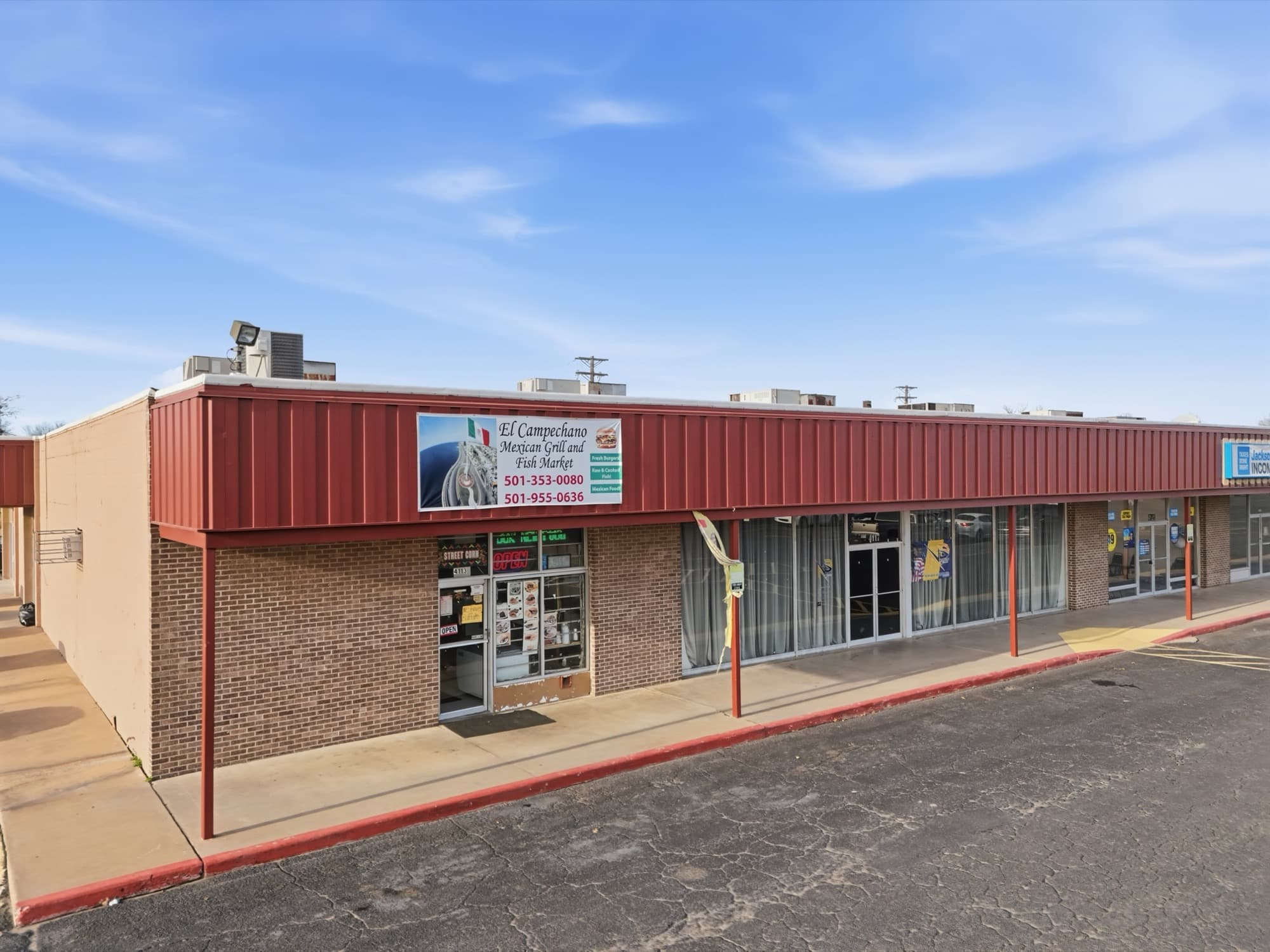 Angled ground level view along the red metal awning of the in line shops at Rose City Center in North Little Rock, Arkansas, with glass storefronts, brick bulkheads and red steel canopy posts receding to the right under a blue sky