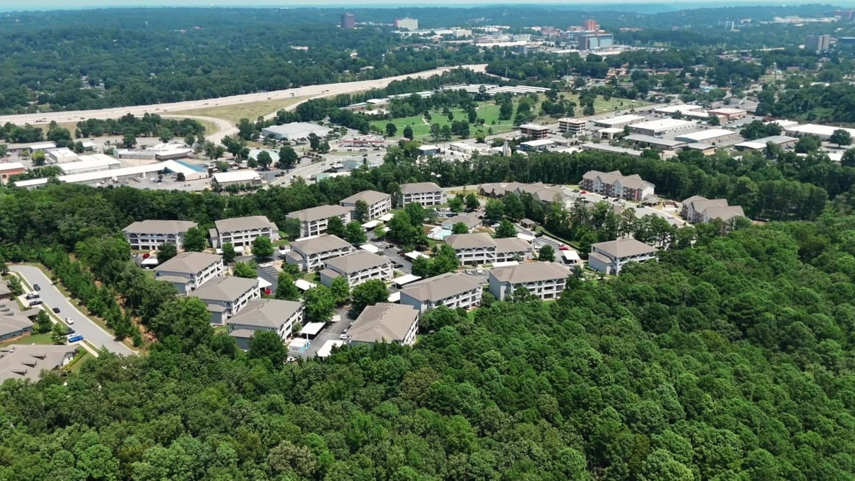 High aerial over an Arkansas apartment community of three story buildings ringed by green tree canopy, with an interstate and a city skyline beyond