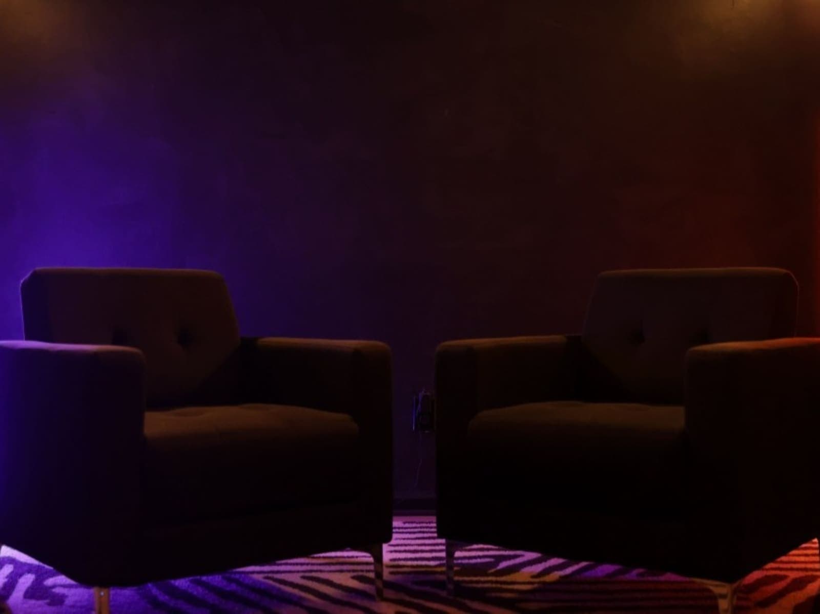 Two dark armchairs facing each other on a patterned rug in a dark walled room, lit purple from the left and orange from the right